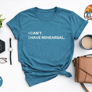 May include: A heather deep teal t-shirt with the text "I CAN'T. I HAVE REHEARSAL." in white. The shirt is styled with a watch, glasses, earrings, a purse, and a cup of coffee.