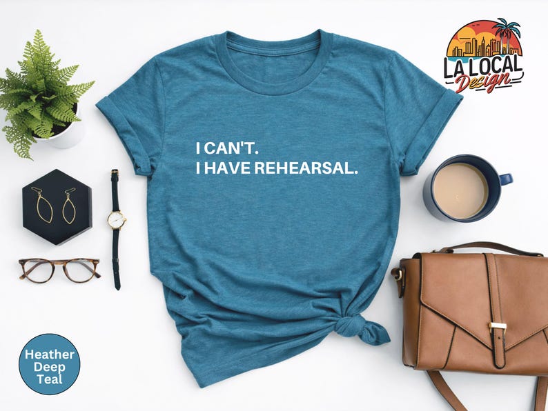 May include: A heather deep teal t-shirt with the text "I CAN'T. I HAVE REHEARSAL." in white. The shirt is styled with a watch, glasses, earrings, a purse, and a cup of coffee.