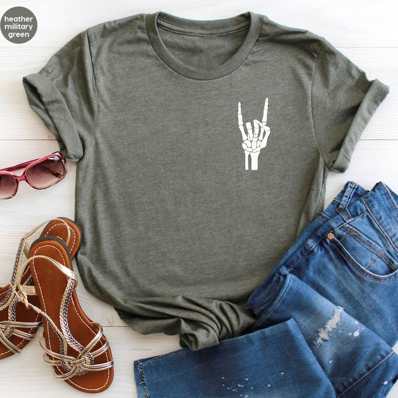 May include: Heather military green t-shirt with a white skeleton hand graphic making a rock and roll gesture. The shirt is paired with blue jeans and brown sandals. The text "heather military green" is in a circle in the upper left corner.