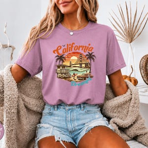 May include: Heather orchid t-shirt with a California Dreamin' graphic. The design features a sunset, palm trees, a lifeguard tower, a car, and the Golden Gate Bridge. The text "California Dreamin'" is in a retro font.