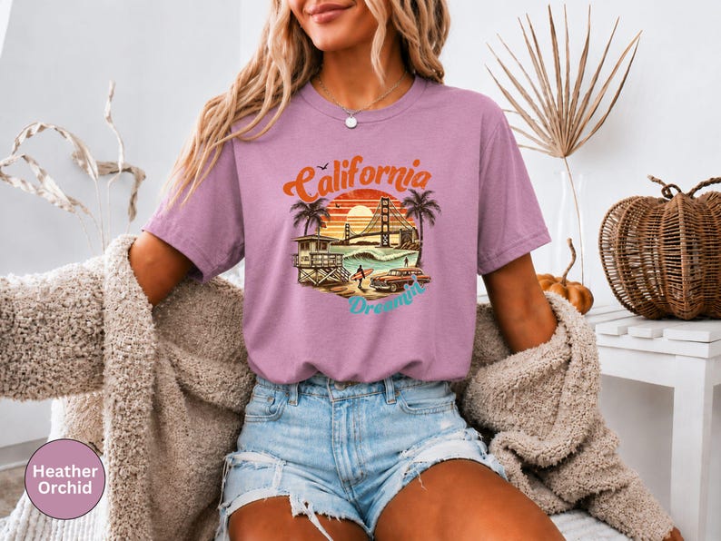 May include: Heather orchid t-shirt with a California Dreamin' graphic. The design features a sunset, palm trees, a lifeguard tower, a car, and the Golden Gate Bridge. The text "California Dreamin'" is in a retro font.