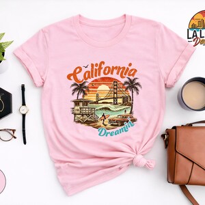 May include: Light pink t-shirt featuring a graphic of a California beach scene with the Golden Gate Bridge, palm trees, and the words "California Dreamin'". The shirt has rolled-up sleeves and a knotted front.