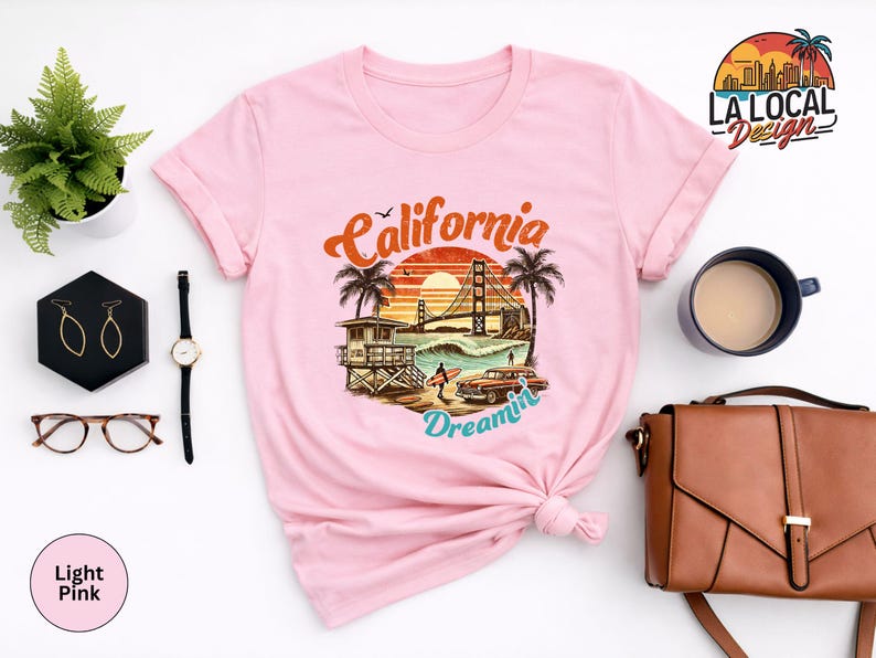 May include: Light pink t-shirt featuring a graphic of a California beach scene with the Golden Gate Bridge, palm trees, and the words "California Dreamin'". The shirt has rolled-up sleeves and a knotted front.