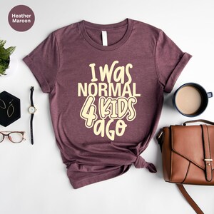 Może przedstawiać: Koszulka w kolorze wrzosowym z kremowym napisem "I Was Normal 4 Kids Ago". Koszulka jest stylizowana z dodatkami, takimi jak zegarek, kolczyki i brązowa torebka. Widoczna jest r&oacute;wnież filiżanka kawy i mała roślina.
