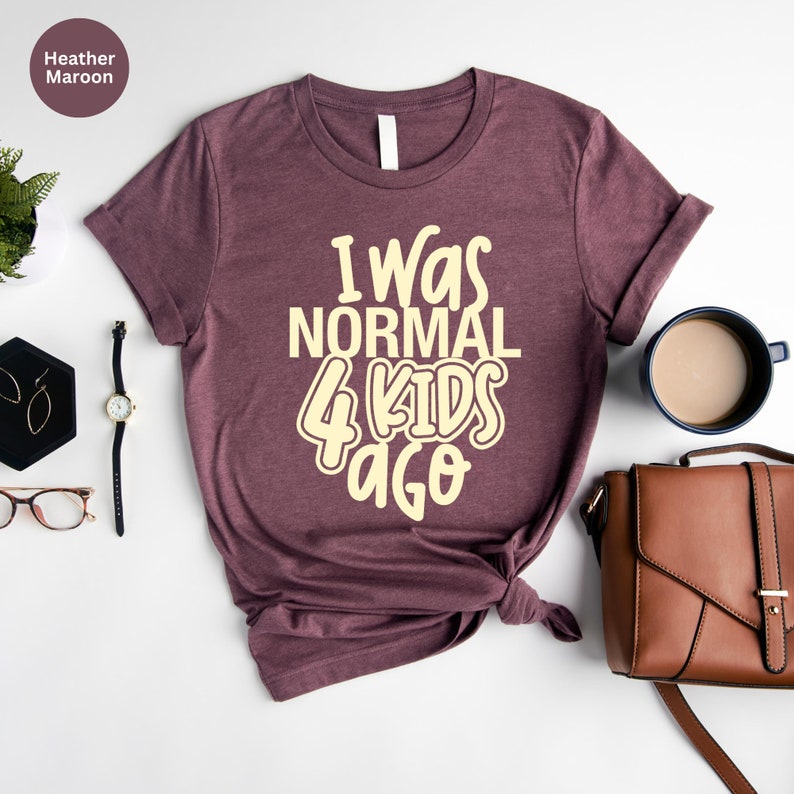 Może przedstawiać: Koszulka w kolorze wrzosowym z kremowym napisem "I Was Normal 4 Kids Ago". Koszulka jest stylizowana z dodatkami, takimi jak zegarek, kolczyki i brązowa torebka. Widoczna jest r&oacute;wnież filiżanka kawy i mała roślina.