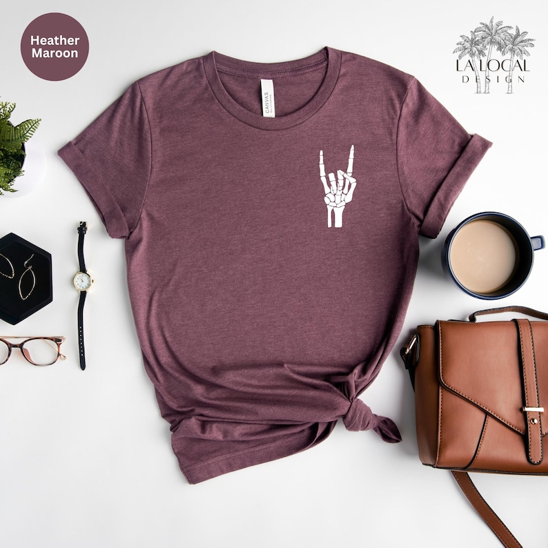 K&ouml;nnte beinhalten: Burgunderfarbenes T-Shirt mit wei&szlig;em Skelett-Hand-Motiv, das das Rock'n'Roll-Zeichen zeigt. Das Shirt wird mit Accessoires wie einer Uhr, Ohrringen und einer braunen Handtasche pr&auml;sentiert. Der Text "Heather Maroon" ist sichtbar.