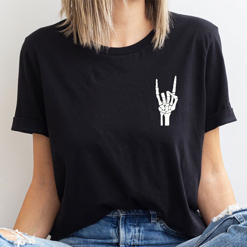 May include: Black t-shirt featuring a small, white skeleton hand graphic making a rock-and-roll hand gesture. The shirt has short sleeves and a relaxed fit.  The image shows the shirt on a model, highlighting its casual style and the subtle yet eye-catching design. The graphic is simple and stylish, suitable for fans of rock music and alternative fashion.