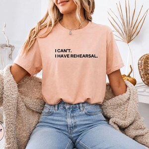 May include: Heather peach-colored t-shirt with the text "I can't. I have rehearsal." in black. The shirt is paired with blue jeans and a beige cardigan. The image also includes decorative elements like a pumpkin and dried plants.