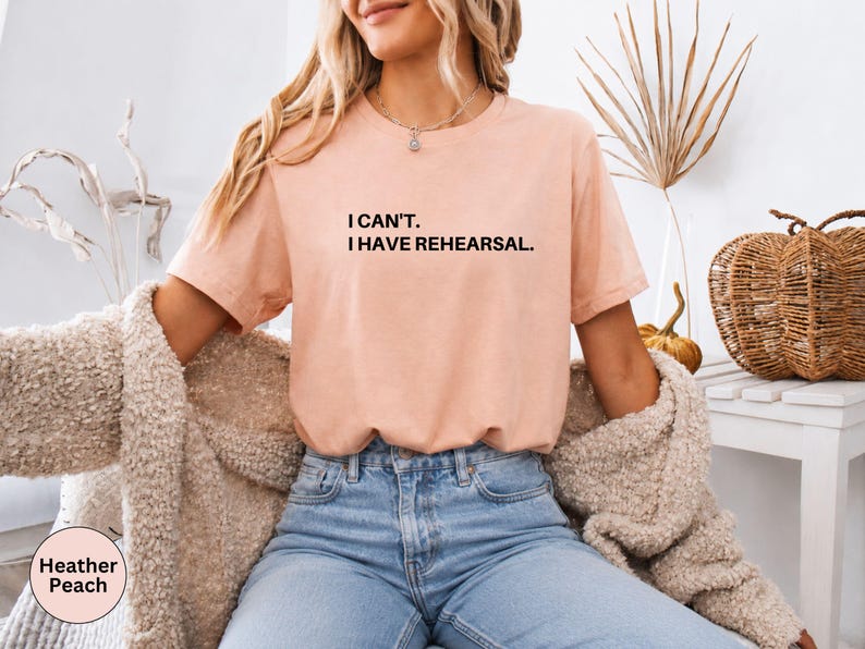 May include: Heather peach-colored t-shirt with the text "I can't. I have rehearsal." in black. The shirt is paired with blue jeans and a beige cardigan. The image also includes decorative elements like a pumpkin and dried plants.