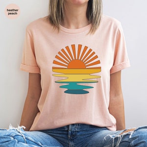 May include: Heather peach t-shirt featuring a retro sunset design. The graphic shows a sun with orange rays above a layered reflection in shades of yellow, teal, and blue. The shirt has rolled-up sleeves and the text "heather peach" is visible.