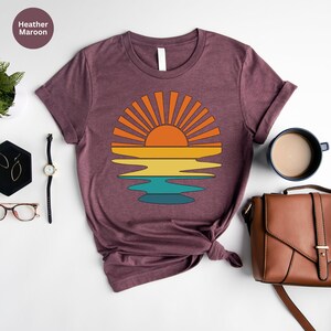 May include: Heather maroon t-shirt featuring a retro sunset design. The graphic shows a sun with orange rays above a layered horizon of yellow, teal, and blue. The shirt is styled with accessories including a watch, earrings, and a purse.
