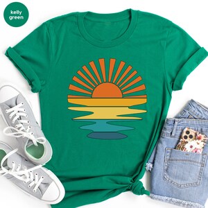 May include: Kelly green t-shirt featuring a retro sunset design. The graphic includes an orange sun with radiating lines, above yellow, and blue horizontal bands, suggesting water. The shirt is paired with gray sneakers and denim shorts.