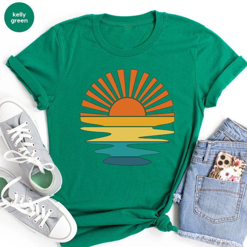 May include: Kelly green t-shirt featuring a retro sunset design. The graphic includes an orange sun with radiating lines, above yellow, and blue horizontal bands, suggesting water. The shirt is paired with gray sneakers and denim shorts.
