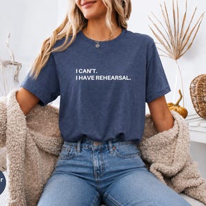 May include: Heather navy blue t-shirt with the text "I can't. I have rehearsal." The shirt is a crew neck style. The model is wearing blue jeans and a beige cardigan. The shirt is a heather navy color.