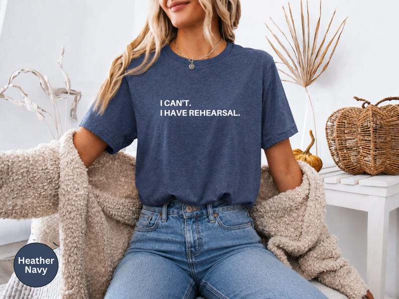 May include: Heather navy blue t-shirt with the text "I can't. I have rehearsal." The shirt is a crew neck style. The model is wearing blue jeans and a beige cardigan. The shirt is a heather navy color.