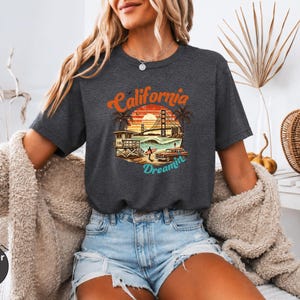 May include: Heather dark grey t-shirt with a colorful graphic of a California beach scene, including the Golden Gate Bridge, palm trees, and a vintage car. The text "California Dreamin'" is arched above the image.