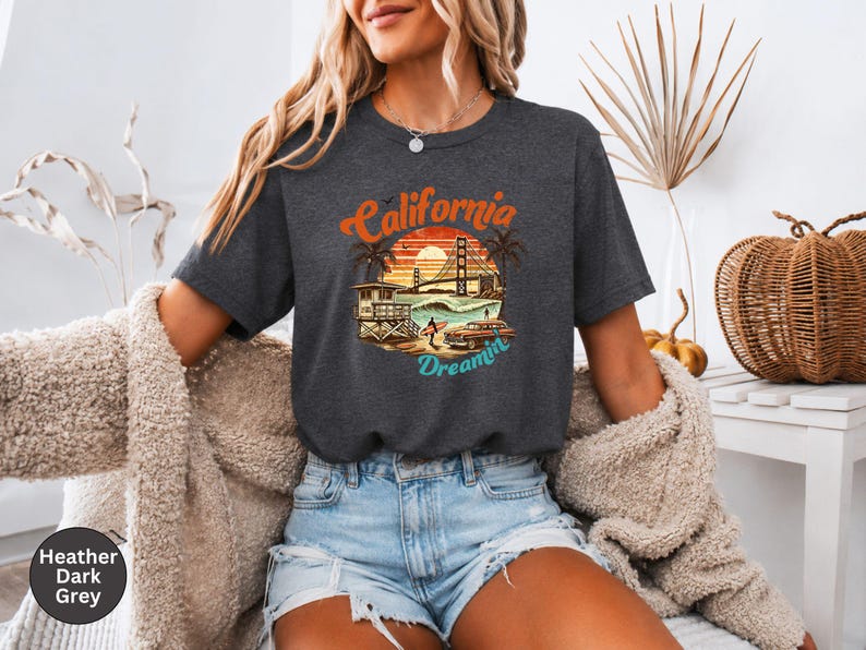 May include: Heather dark grey t-shirt with a colorful graphic of a California beach scene, including the Golden Gate Bridge, palm trees, and a vintage car. The text "California Dreamin'" is arched above the image.