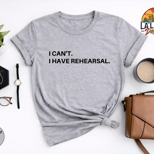 May include: Heather light grey t-shirt with the text "I CAN'T. I HAVE REHEARSAL." A black watch, gold earrings, and a brown purse are next to the shirt. A small plant and a cup of coffee are also in the image.
