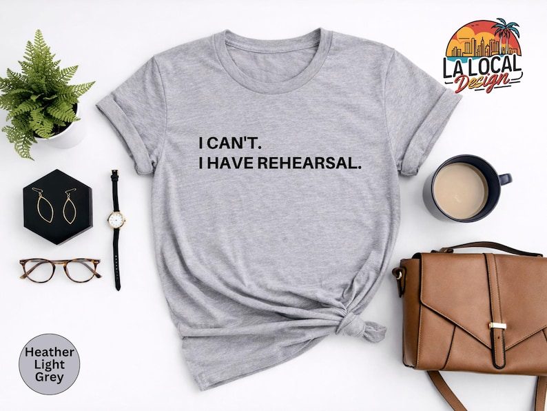 May include: Heather light grey t-shirt with the text "I CAN'T. I HAVE REHEARSAL." A black watch, gold earrings, and a brown purse are next to the shirt. A small plant and a cup of coffee are also in the image.