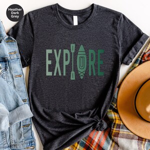 Puede incluir: Camiseta gris oscuro jaspeado con la palabra "EXPLORE" en letras verdes. La "O" se reemplaza con un gr&aacute;fico de kayak y remos. La camiseta se combina con una chaqueta vaquera, una bufanda a cuadros y un sombrero marr&oacute;n.