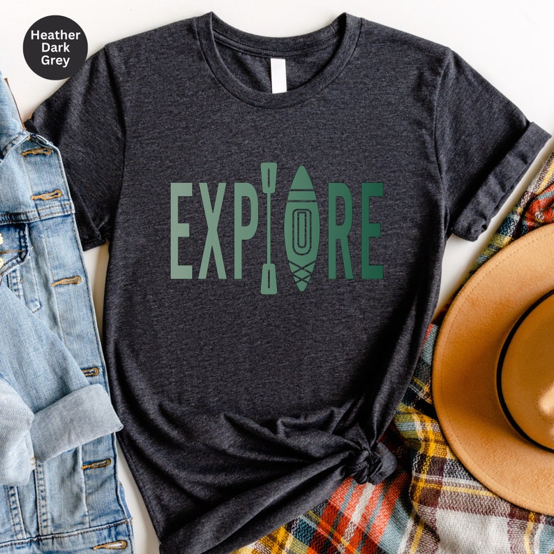 Puede incluir: Camiseta gris oscuro jaspeado con la palabra "EXPLORE" en letras verdes. La "O" se reemplaza con un gr&aacute;fico de kayak y remos. La camiseta se combina con una chaqueta vaquera, una bufanda a cuadros y un sombrero marr&oacute;n.