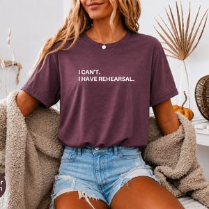 May include: Heather maroon t-shirt with the text "I CAN'T. I HAVE REHEARSAL." The shirt is paired with denim shorts and a beige cardigan. A decorative pumpkin and woven basket are in the background.