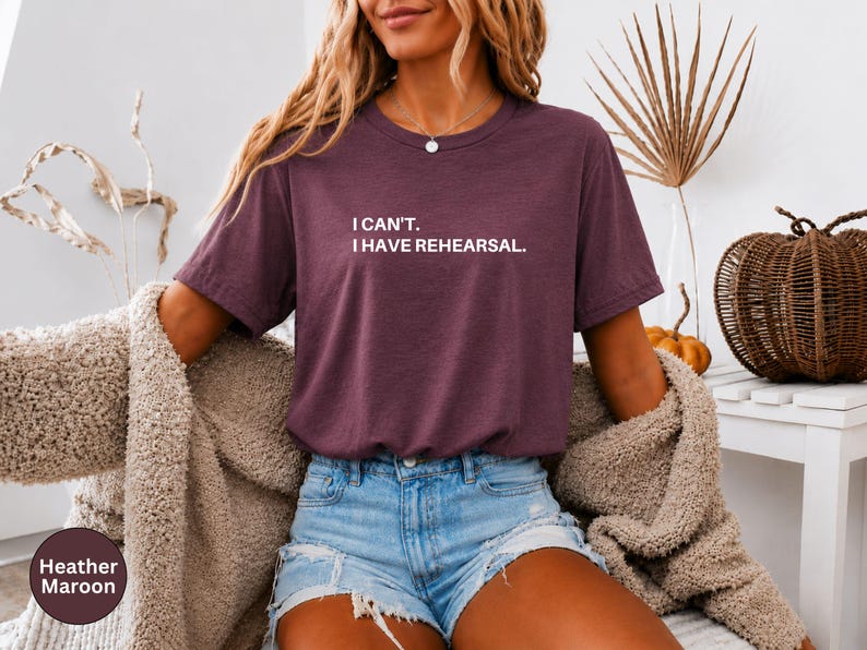 May include: Heather maroon t-shirt with the text "I CAN'T. I HAVE REHEARSAL." The shirt is paired with denim shorts and a beige cardigan. A decorative pumpkin and woven basket are in the background.