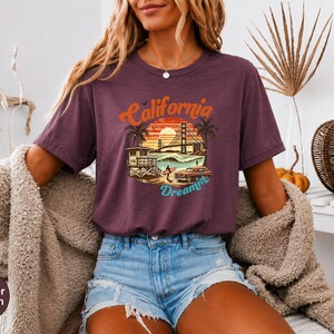 May include: Heather maroon t-shirt with a graphic that says "California Dreamin'". The design features a sunset, palm trees, the Golden Gate Bridge, and a vintage car. The shirt is paired with denim shorts.