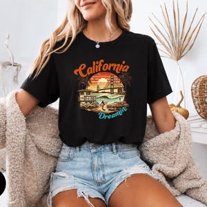 May include: Black t-shirt with a colorful graphic featuring the words "California Dreamin'" above an image of the Golden Gate Bridge, palm trees, and a lifeguard station. The shirt is paired with denim shorts.