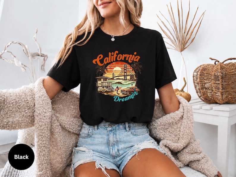 May include: Black t-shirt with a colorful graphic featuring the words "California Dreamin'" above an image of the Golden Gate Bridge, palm trees, and a lifeguard station. The shirt is paired with denim shorts.