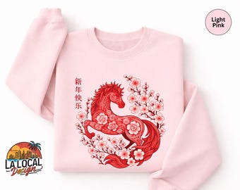 May include: Light pink sweatshirt featuring a red horse design surrounded by cherry blossoms. The horse is intricately detailed with floral patterns. The text "新年快乐" is printed vertically on the left side. The sweatshirt has a crew neck and long sleeves.