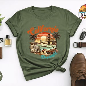 May include: Military green t-shirt featuring a California Dreamin' graphic with a sunset, palm trees, and the Golden Gate Bridge. The shirt is styled with accessories like glasses, a wallet, and brown leather shoes.