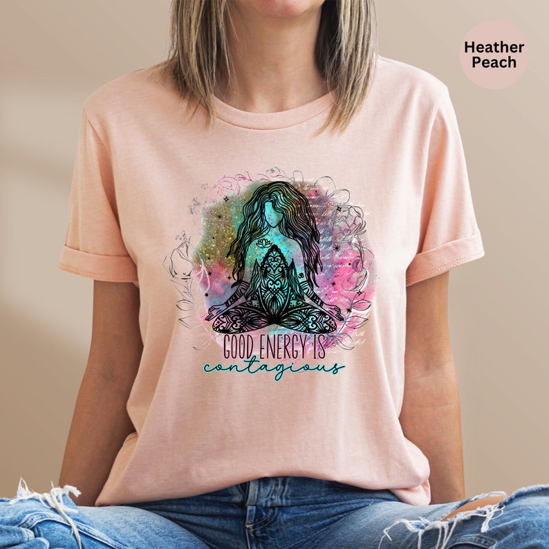 Peut inclure: T-shirt p&ecirc;che clair avec un motif de femme assise en position de yoga. Le graphisme comprend une femme aux longs cheveux, des motifs complexes et le texte turquoise "GOOD ENERGY IS contagious". Le design est entour&eacute; d'&eacute;l&eacute;ments floraux et aquarelles.