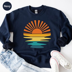 May include: Navy blue sweatshirt featuring a retro sunset design. The graphic shows an orange sun with yellow, teal, and orange stripes, reflecting on the water. The sweatshirt is paired with blue jeans and white sneakers.