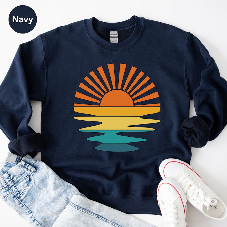May include: Navy blue sweatshirt featuring a retro sunset design. The graphic shows an orange sun with yellow, teal, and orange stripes, reflecting on the water. The sweatshirt is paired with blue jeans and white sneakers.