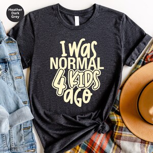 Może przedstawiać: Ciemnoszara koszulka z napisem "I Was Normal 4 Kids Ago" w zabawnym, jasnoż&oacute;łtym kroju pisma. Koszulka z okrągłym dekoltem jest prezentowana na białej powierzchni z kurtką dżinsową i kapeluszem.