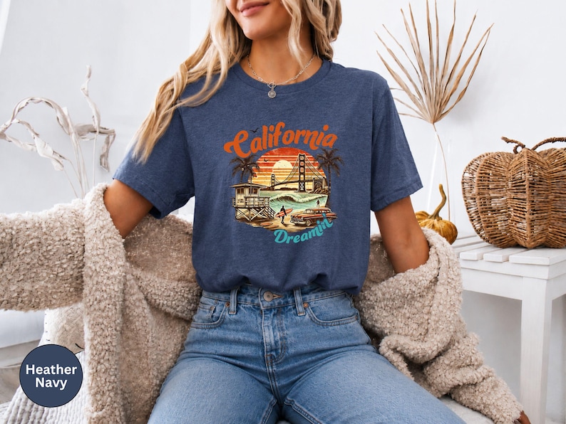 May include: Heather navy blue t-shirt with a California Dreamin' graphic. The design features a sunset, palm trees, the Golden Gate Bridge, and a vintage car. The shirt is a relaxed fit and made of soft material.