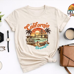 May include: Sand-colored t-shirt featuring a California-themed graphic with the Golden Gate Bridge, palm trees, and the words "California Dreamin'". The design includes a vintage car and a lifeguard tower. Accessories include a watch, earrings, and a handbag.
