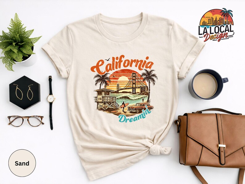 May include: Sand-colored t-shirt featuring a California-themed graphic with the Golden Gate Bridge, palm trees, and the words "California Dreamin'". The design includes a vintage car and a lifeguard tower. Accessories include a watch, earrings, and a handbag.