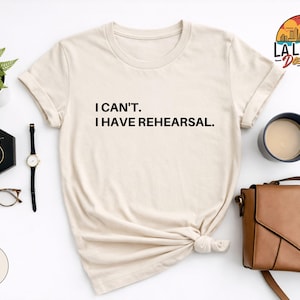 May include: Sand-colored t-shirt with the text "I CAN'T. I HAVE REHEARSAL." A black watch, gold earrings, and a brown purse are next to the shirt. A small plant and a cup of coffee are also in the image. The shirt is knotted at the waist.