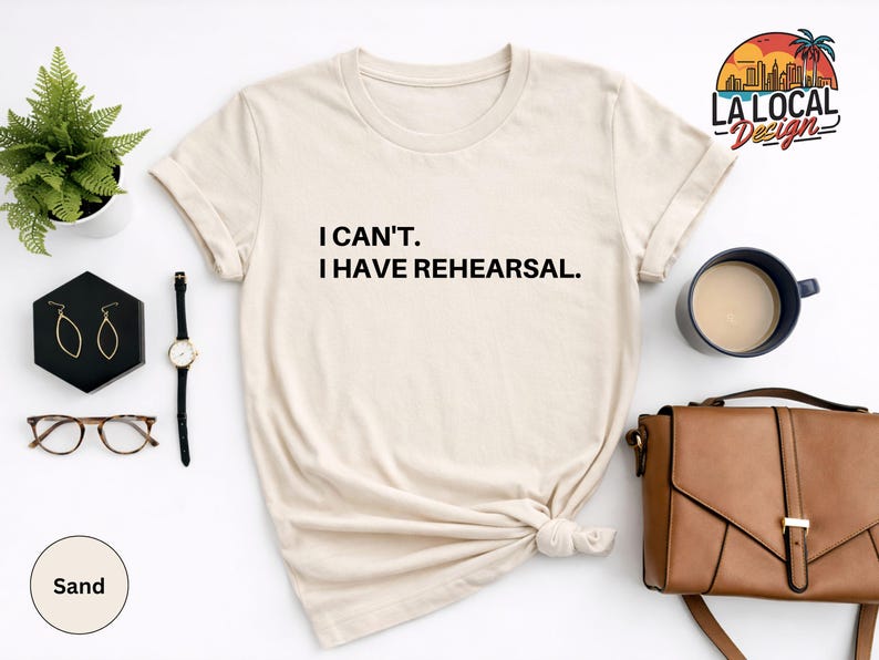 May include: Sand-colored t-shirt with the text "I CAN'T. I HAVE REHEARSAL." A black watch, gold earrings, and a brown purse are next to the shirt. A small plant and a cup of coffee are also in the image. The shirt is knotted at the waist.
