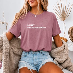 May include: Heather mauve t-shirt with the text "I can't. I have rehearsal." The shirt is a short-sleeved crew neck. The model is wearing denim shorts and a beige cardigan. A decorative pumpkin and woven basket are in the background.