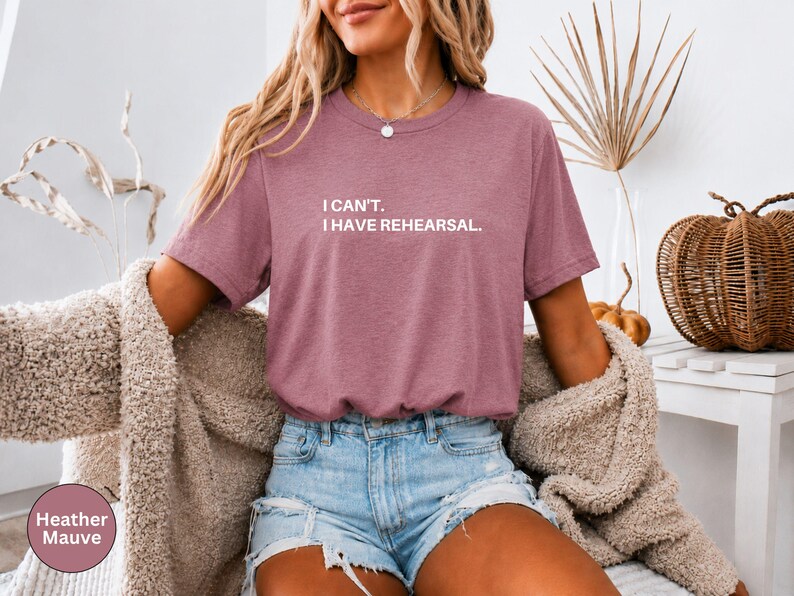 May include: Heather mauve t-shirt with the text "I can't. I have rehearsal." The shirt is a short-sleeved crew neck. The model is wearing denim shorts and a beige cardigan. A decorative pumpkin and woven basket are in the background.