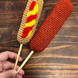 Peut inclure: Deux hot-dogs au crochet sur des bâtonnets en bois. L'un est beige avec des détails jaunes et rouges, ressemblant à de la moutarde et du ketchup. L'autre est de couleur rouge-orangé uni. Les articles sont présentés sur une surface en bois foncé.