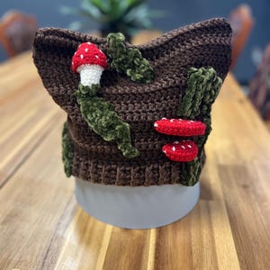 Op de afbeelding: Een bruine gehaakte hoed met rode en witte paddenstoelen en groene bladeren. De hoed is gemaakt van zacht, getextureerd garen. Een uniek, handgemaakt accessoire dat op het hoofd gedragen kan worden.