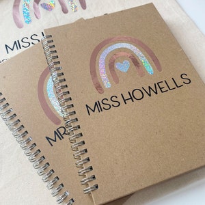 May include: Two brown spiral notebooks with a holographic rainbow and heart design. The notebooks have the text "Miss Howells" and "Mr." printed on them.