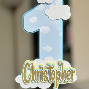 May include: A blue number one cake topper with white clouds and the name Christopher in gold glitter letters on a white cloud.