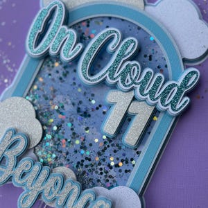 May include: A layered cake topper with a cloud and rainbow theme. The topper features the words "On Cloud 11" in glittery teal and silver. The name "Beyonca" is also included. The design incorporates blue, white, and silver elements, with a purple background.