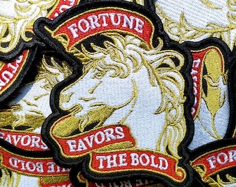 Op de afbeelding: Een verzameling geborduurde patches met een witte paardenkop in profiel, omlijnd in zwart. Elke patch bevat een rood lint met de witte tekst FORTUNE boven het paard en FAVORS THE BOLD onder het paard in goudkleurige letters.