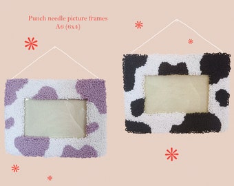 May include: Two punch needle picture frames, A6 size (15x10 cm), with a cow-like pattern. One frame has white and lavender spots, the other has white and black spots. Each frame is suspended by a white string.